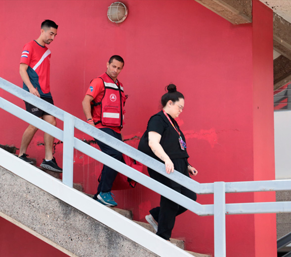 Se muestran dos hombres y una mujer bajando por las gradas de seguridad del edificio. Uno de los hombres porta un chaleco de la  brigada de evacuación. 